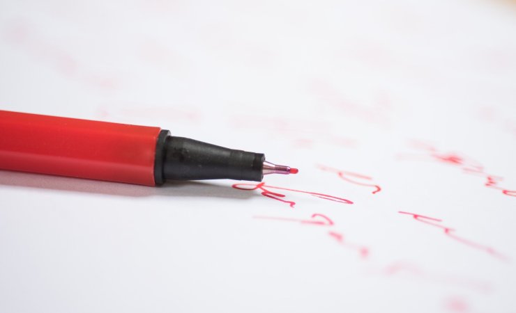 Image d'un stylo rouge posé sur une feuille où l'on aperçoit une partie de texte écrit à la main en rouge.